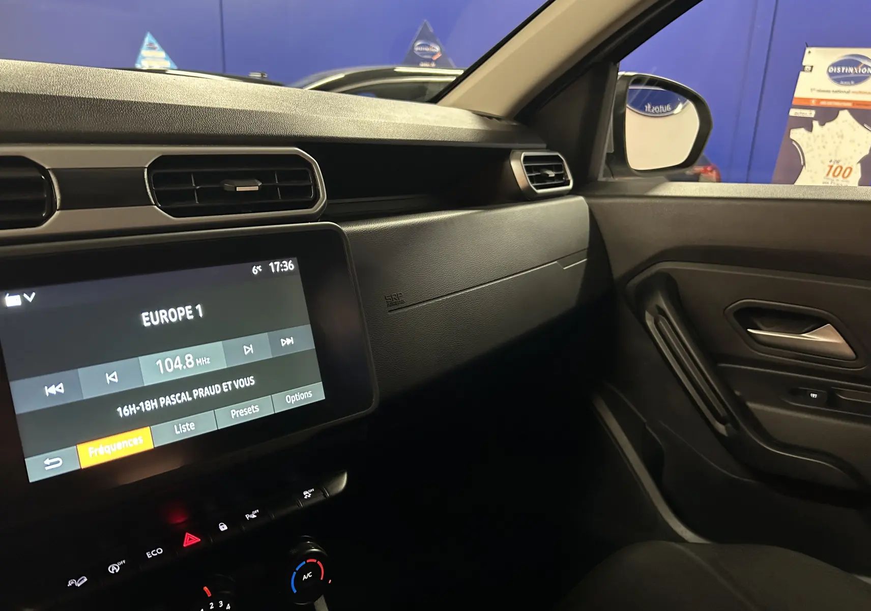 Vue intérieure côté passager du tableau de bord noir du Dacia Duster 2024 avec écran tactile et commandes de climatisation.