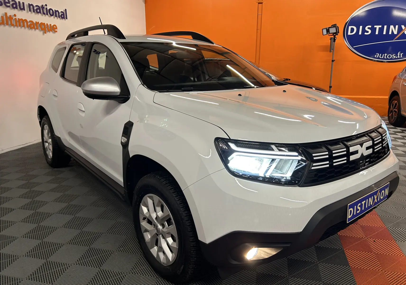 Vue 3/4 avant d'un Dacia Duster blanc 2024 avec phares allumés et calandre noire distinctive dans un showroom.