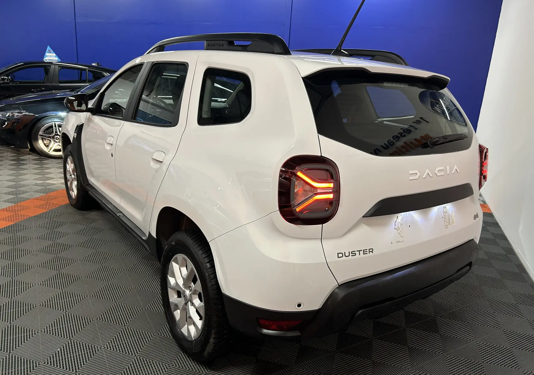Vue 3/4 arrière droite d'un Dacia Duster blanc 2024 avec feux arrière allumés et barres de toit noires.