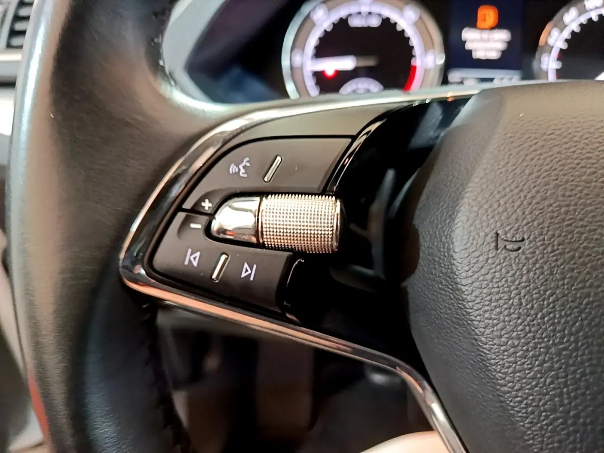 Gros plan sur les commandes gauche du volant cuir noir de la Skoda Superb blanche, avec tableau de bord numérique en arrière-plan.