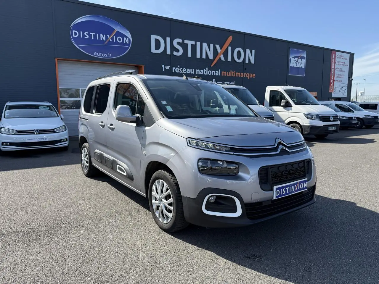Vue 3/4 avant d'un Citroën Berlingo gris foncé garé devant un concessionnaire Distinxion par temps clair.