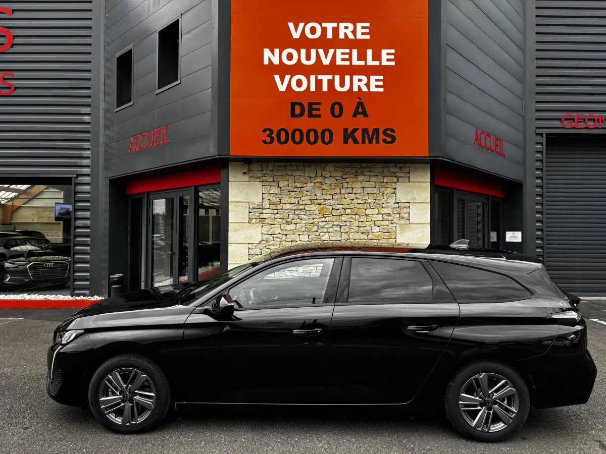 Profil latéral gauche d'une Peugeot 308 noire 2024 garée devant un bâtiment avec panneau orange "Votre nouvelle voiture"