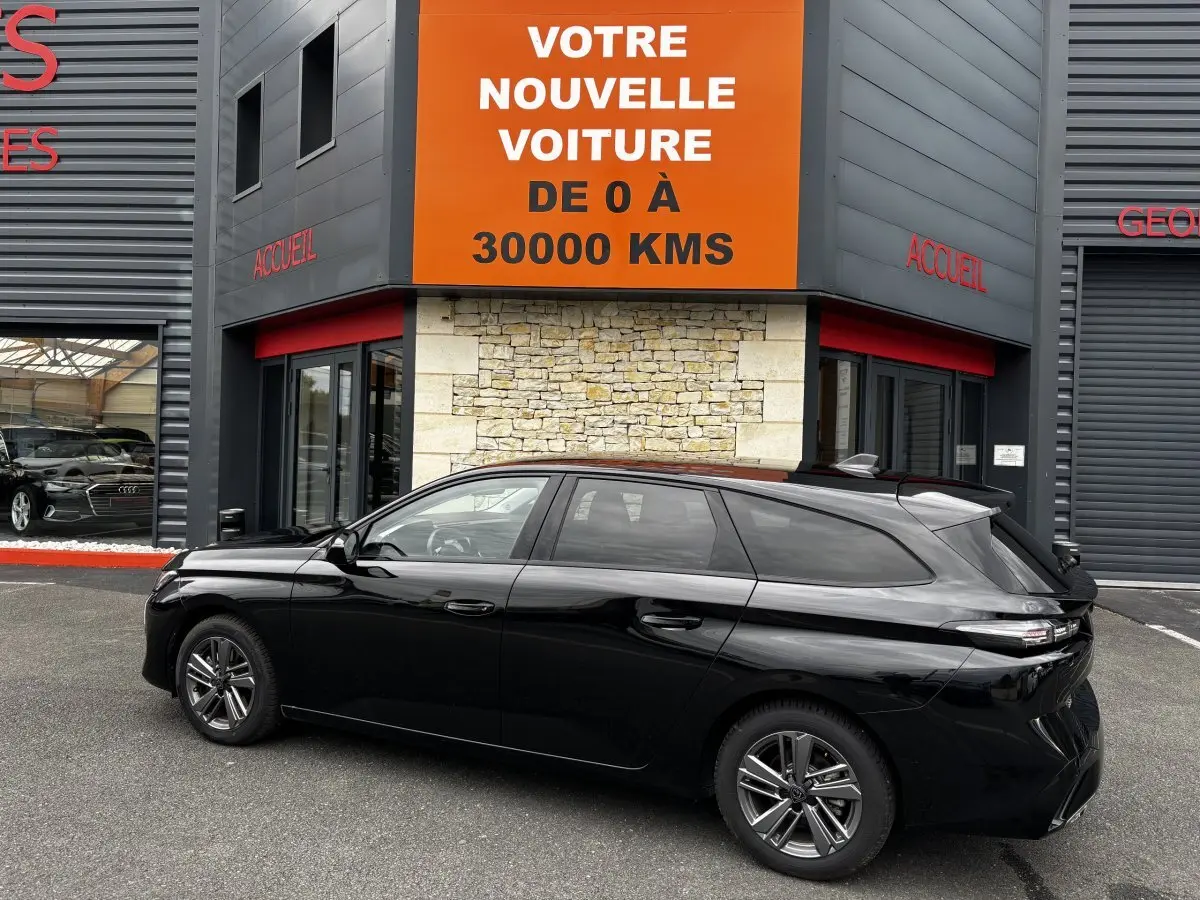 Peugeot 308 PureTech 130 noire vue de profil côté gauche, stationnée devant un bâtiment avec panneau orange.