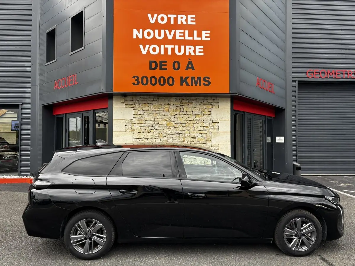 Profil côté gauche d'une Peugeot 308 noire 2024 garée devant un bâtiment avec panneau orange "Votre nouvelle voiture".