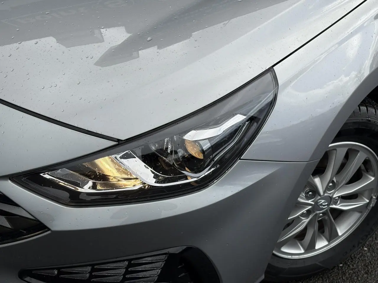 Gros plan sur le phare avant droit et la jante alliage gris argent d'une Hyundai i30 SW T-GDI 2025 sous la pluie.
