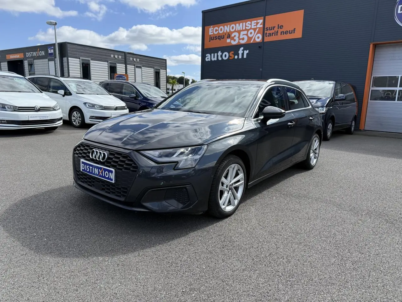 Audi A3 Sportback gris Manhattan métallisé vue 3/4 avant droit sur parking avec enseigne orange en arrière-plan