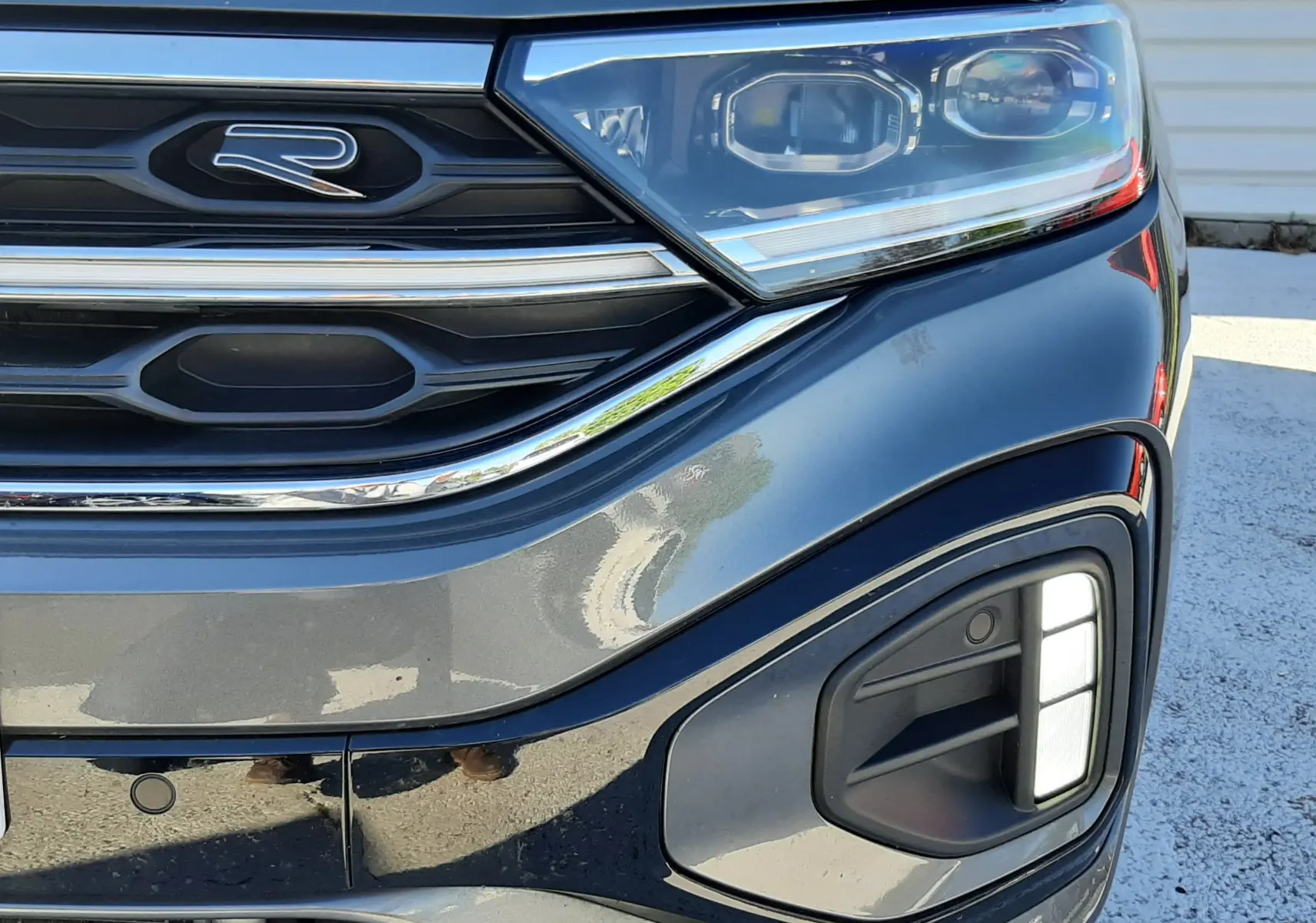 Gros plan sur l'avant droit du Volkswagen T-Roc gris Indium, mettant en valeur le logo R et les phares LED distinctifs.