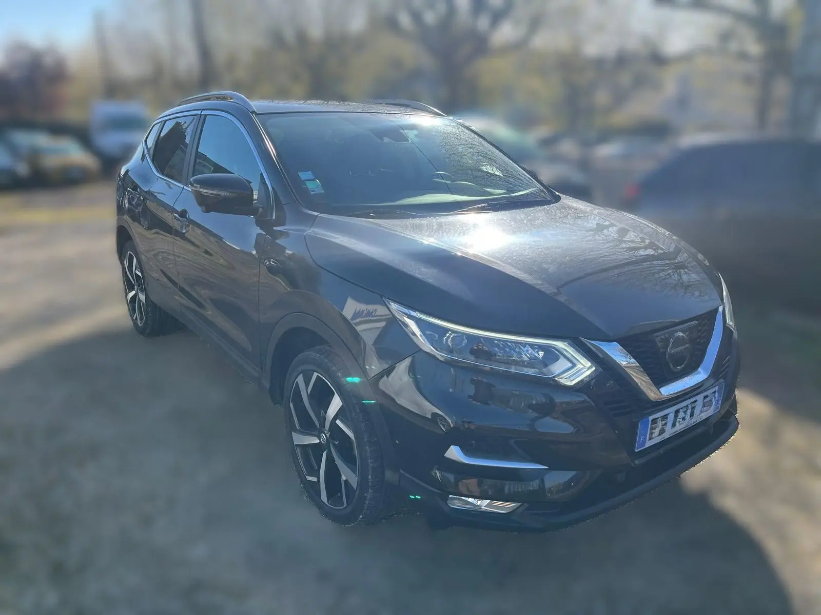 Nissan Qashqai noir en 3/4 avant droit, avec jantes alu et barres de toit visibles au soleil.