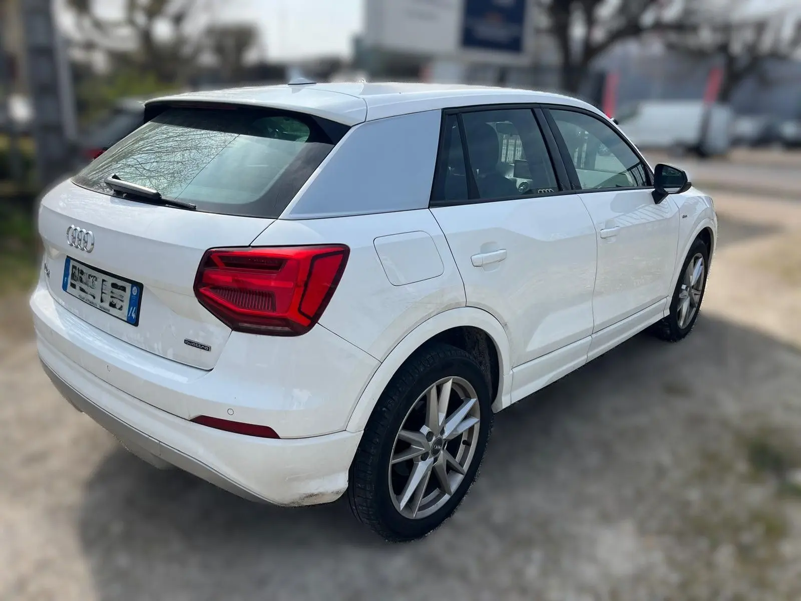 Audi Q2 blanc vue 3/4 arrière droit, avec jantes alu et feux arrière rouges bien visibles.