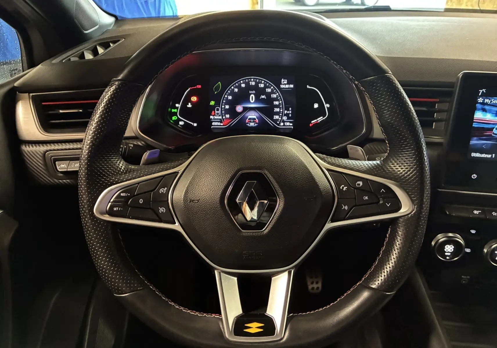 Vue rapprochée du volant noir perforé et du tableau de bord numérique du Renault Captur 1.3 TCE 140 EDC RS Line gris, intérieur moderne.