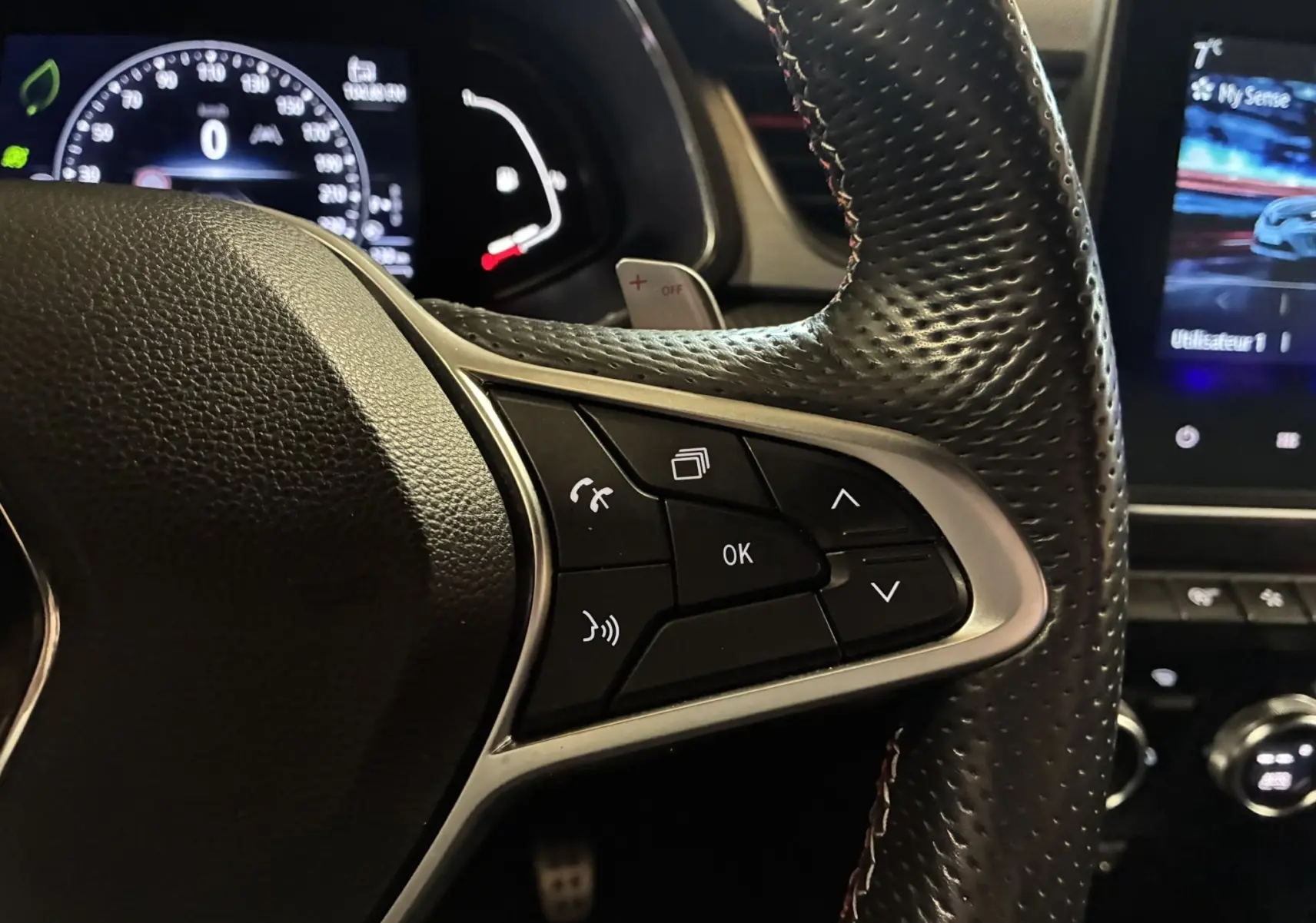 Gros plan sur le côté droit du volant en cuir perforé noir du Renault Captur gris, avec commandes multifonctions et tableau de bord numérique en arrière-plan.