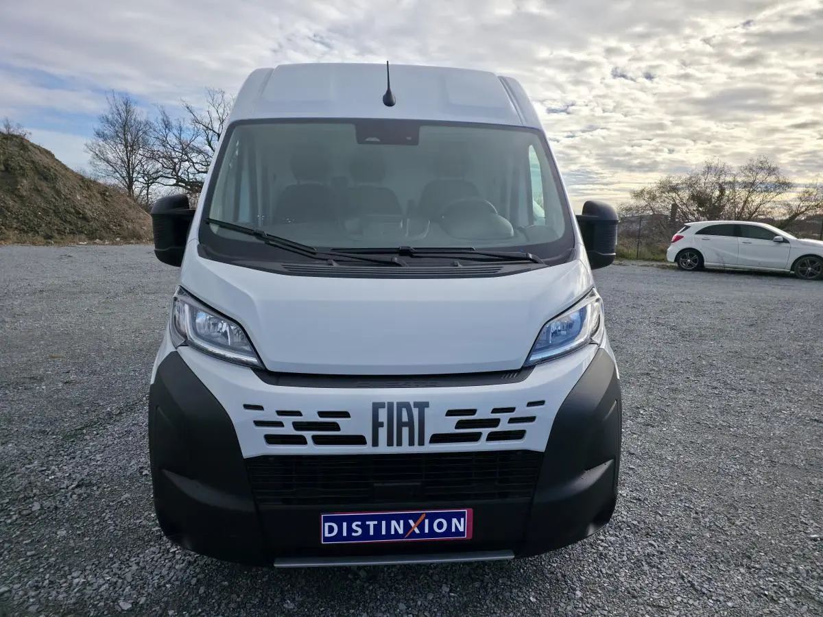 Vue de face d'un FIAT Ducato Fourgon blanc avec pare-chocs noir et logo FIAT bien visible sur un terrain extérieur.
