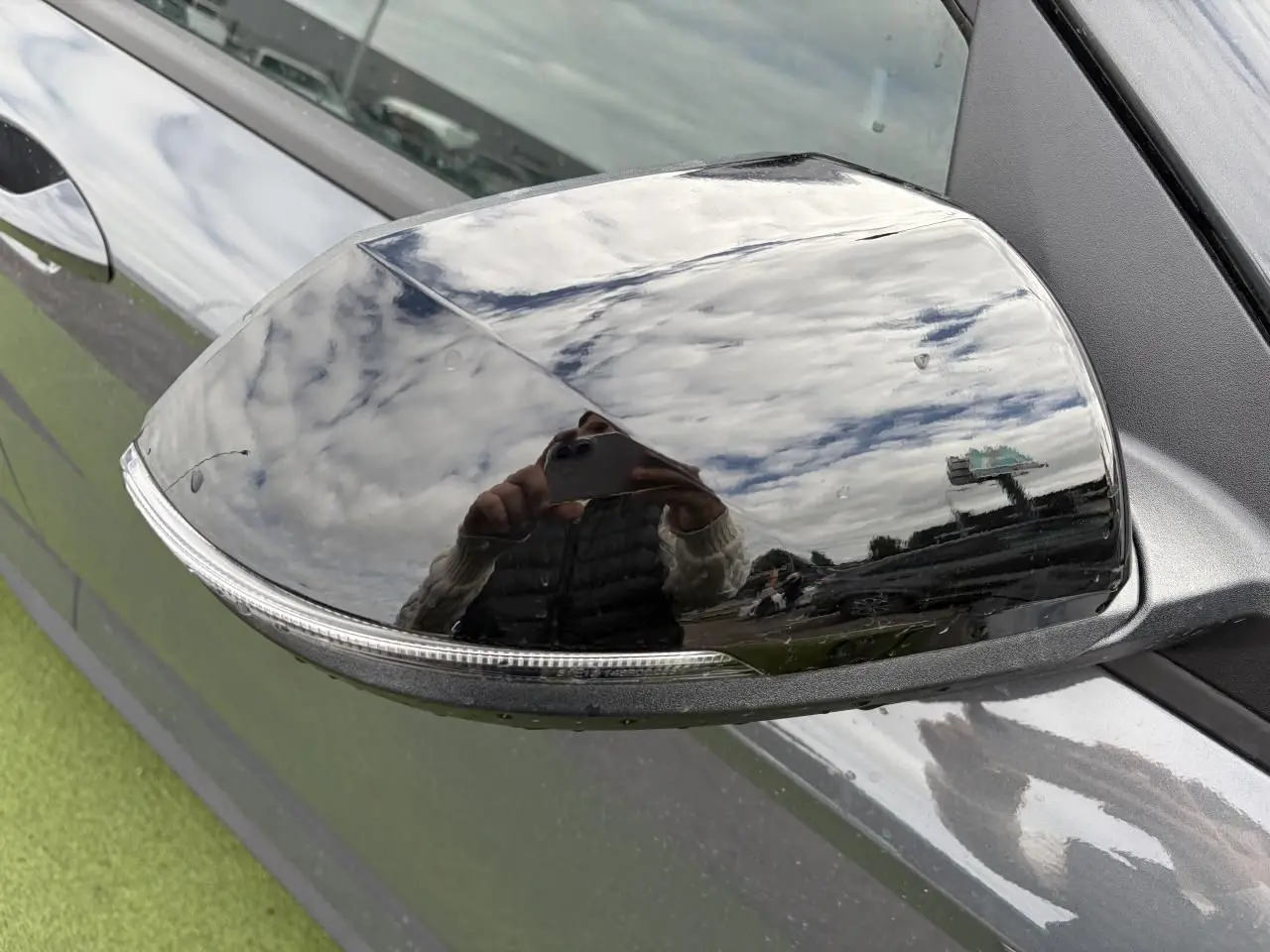 Rétroviseur droit noir brillant du Hyundai Kona II 1.6 Hybrid 138 N Line en Ecotronic Grey, reflet du ciel nuageux visible.