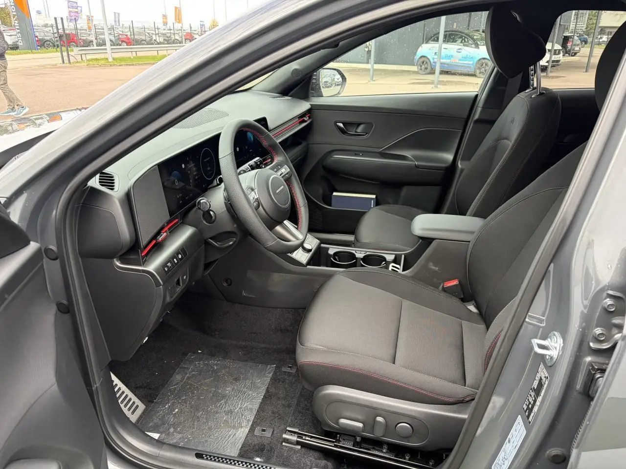 Vue intérieure côté conducteur du Hyundai Kona gris Ecotronic, avec tableau de bord numérique et volant cuir à surpiqûres rouges.