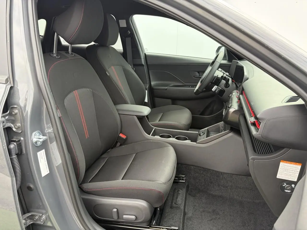 Vue intérieure côté conducteur du Hyundai Kona II 2026 gris Ecotronic, sièges noirs avec surpiqûres rouges et console centrale.