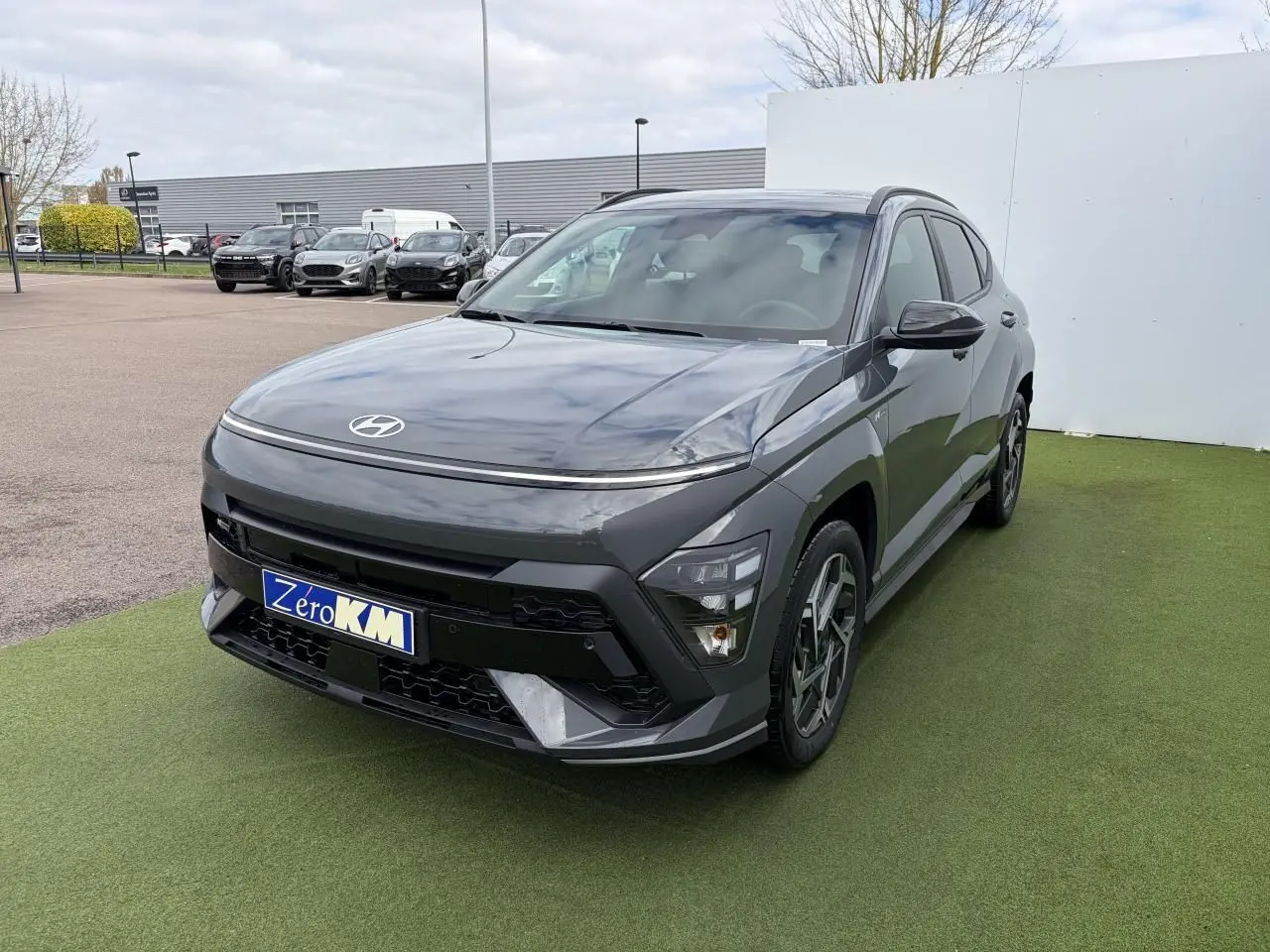 Hyundai Kona II 1.6 Hybrid N Line gris Ecotronic en 3/4 avant droit sur sol vert avec calandre noire distinctive.