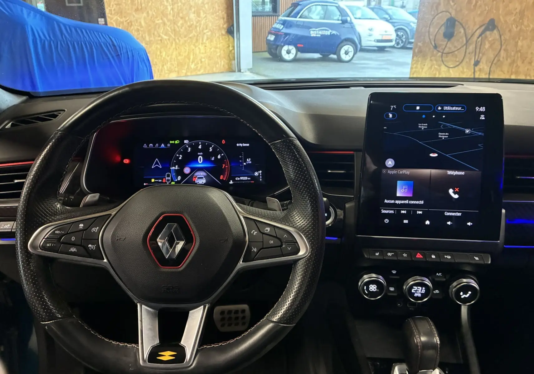 Vue intérieure centrée sur le volant noir perforé avec logo Renault rouge, tableau de bord digital et écran tactile central allumé.