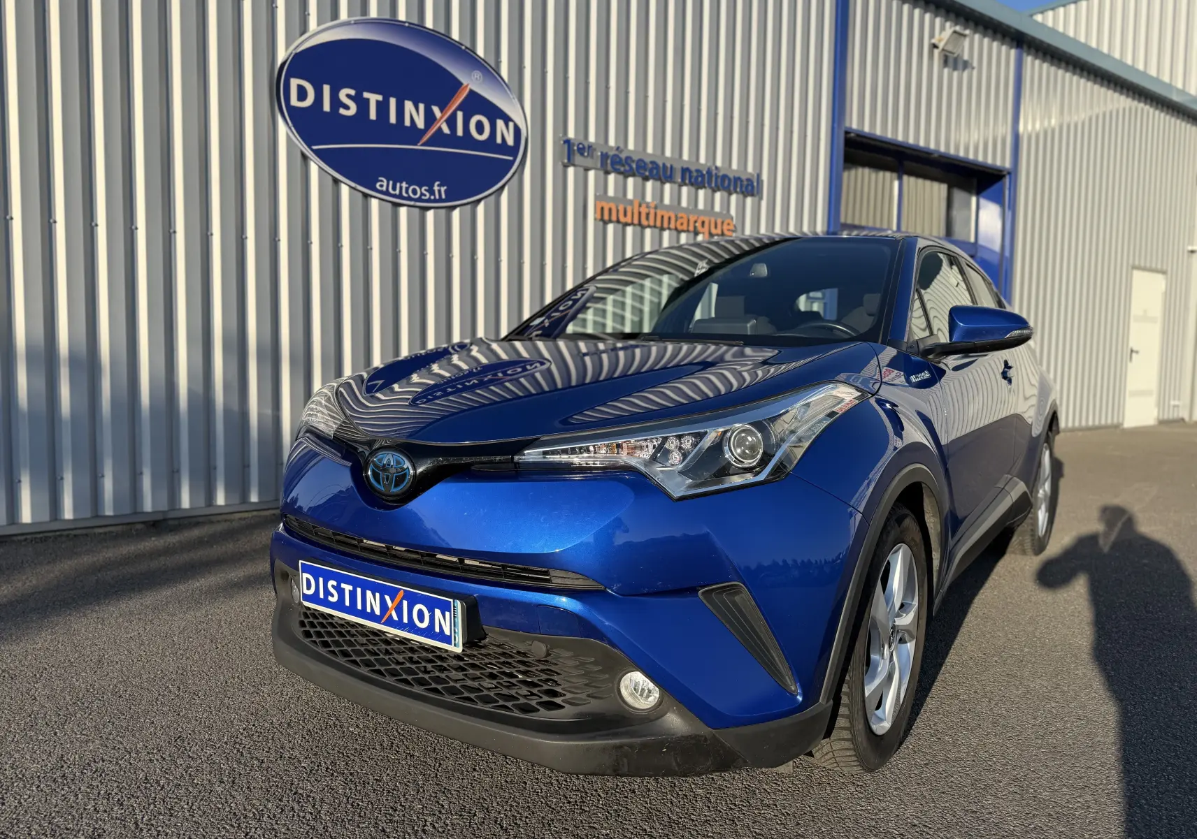 Toyota C-HR bleu Nebula Metal en 3/4 avant droit, avec calandre noire et phares anguleux, stationné devant un bâtiment.
