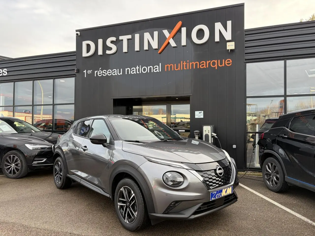 Nissan Juke II 1.6 Hybrid noir métal en 3/4 avant droit, avec jantes alliage et calandre distinctive, devant un showroom.