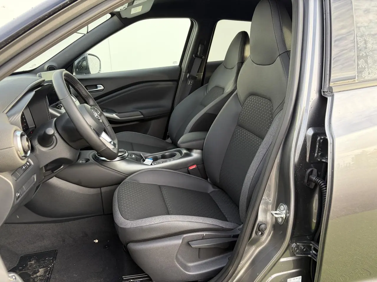 Vue intérieure côté conducteur du Nissan Juke II 1.6 Hybrid 2025, sièges en tissu noir et gris avec console centrale visible.