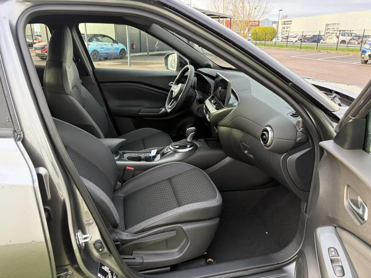 Vue intérieure côté conducteur du Nissan Juke II noir métal, montrant les sièges, volant cuir et console centrale moderne.