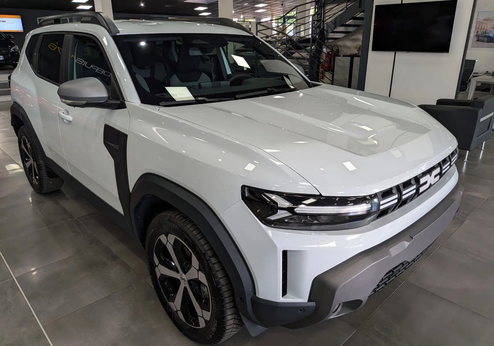 Vue 3/4 avant droite du Dacia Duster Hybrid-G blanc Glacier avec détails noirs contrastants et jantes bi-ton en showroom.