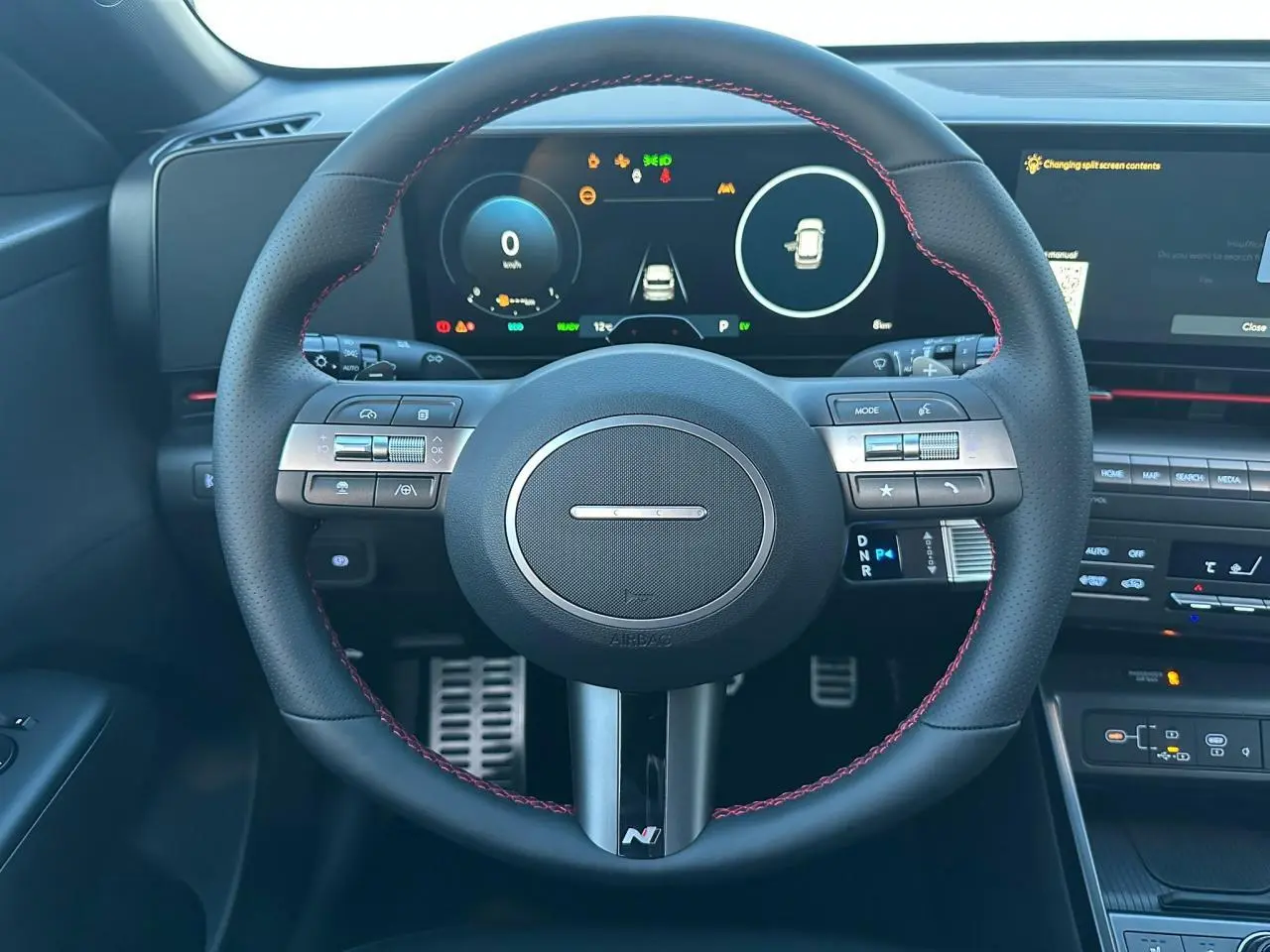 Vue intérieure centrée sur le volant cuir noir avec surpiqûres rouges du Hyundai Kona 2025, tableau de bord numérique visible.