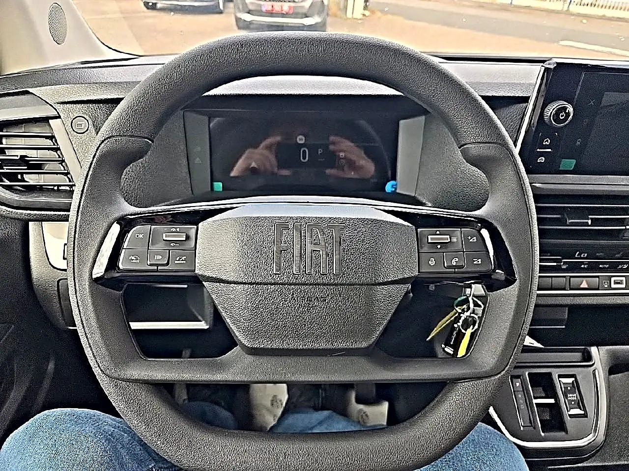 Vue intérieure centrée sur le volant noir FIAT avec commandes intégrées et tableau de bord numérique en arrière-plan.