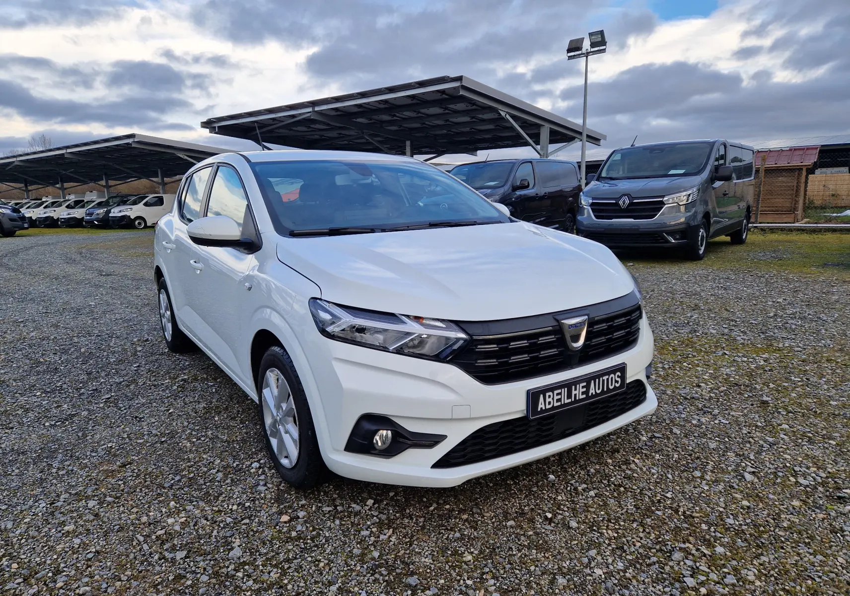 Dacia Sandero blanc vue 3/4 avant droit sur parking gravillonné avec plusieurs véhicules en arrière-plan.