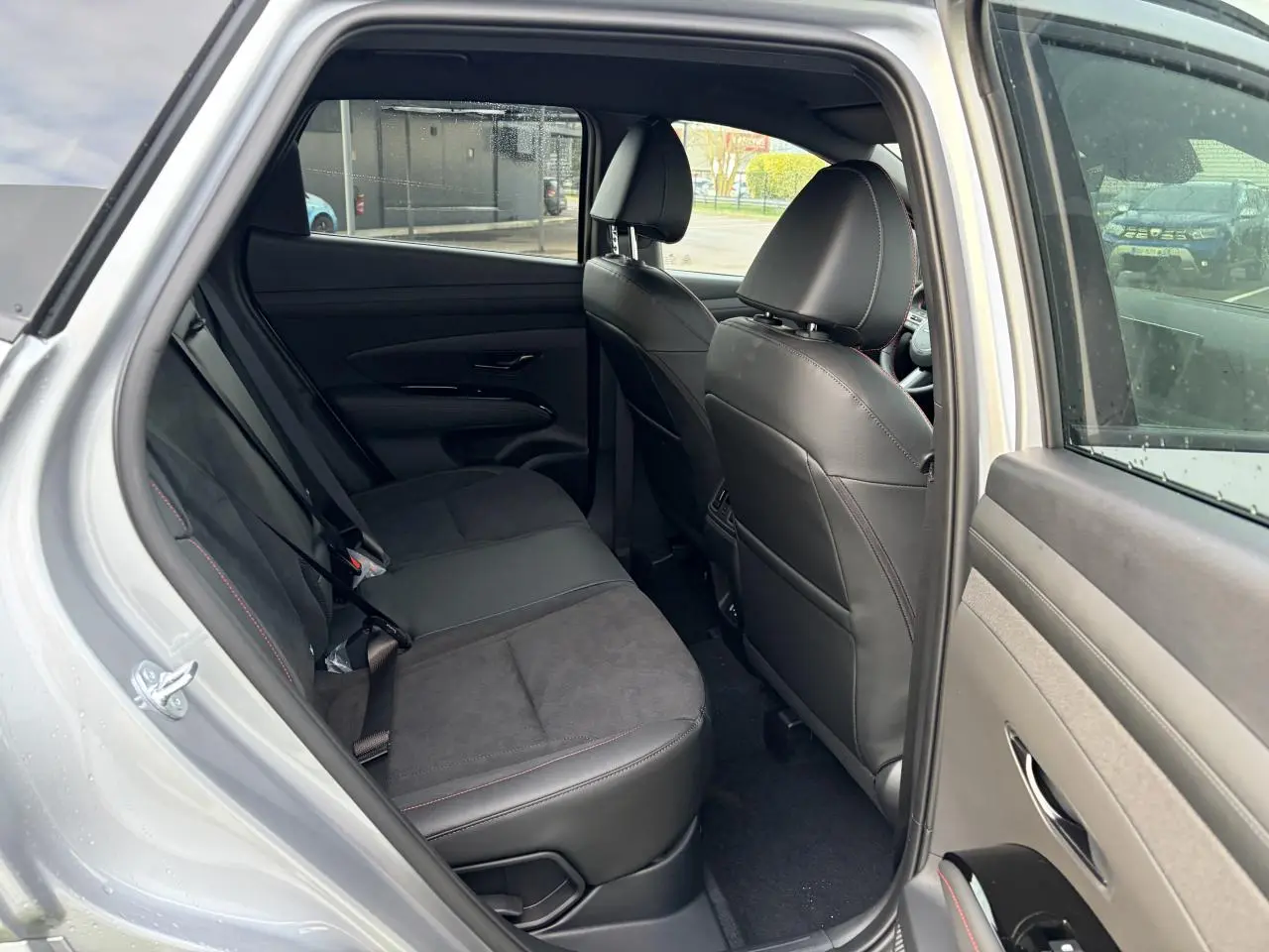Vue intérieure arrière côté gauche du Hyundai Tucson 2025 gris métallisé, avec sellerie cuir/alcantara noire et surpiqûres rouges.