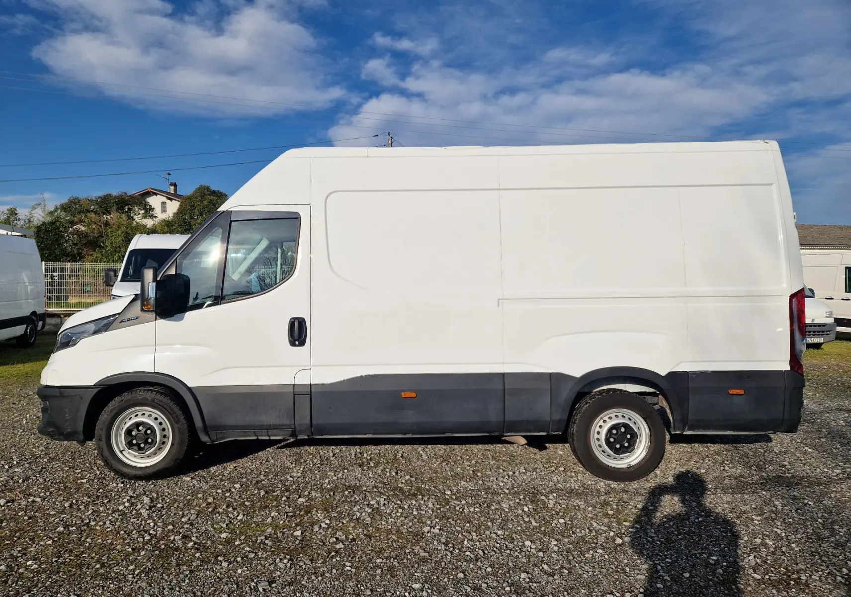 Vue de profil côté gauche d’un Iveco Daily Fourgon blanc 2023 avec bas de caisse noir sur un sol gravillonné.