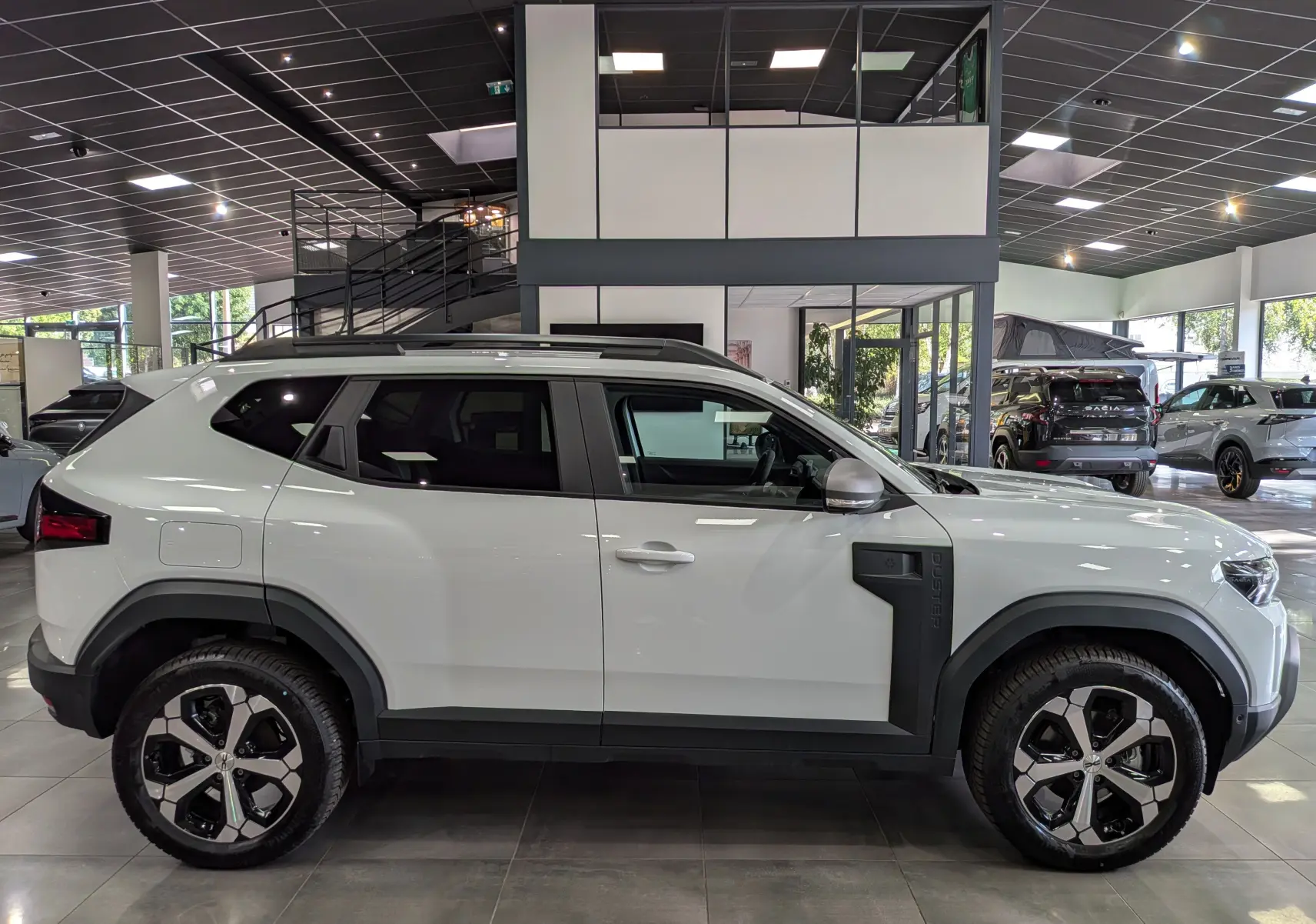 Vue de profil côté gauche d'un Dacia Duster Hybrid-G blanc Glacier en showroom, avec jantes bi-ton et protections noires sur les ailes.