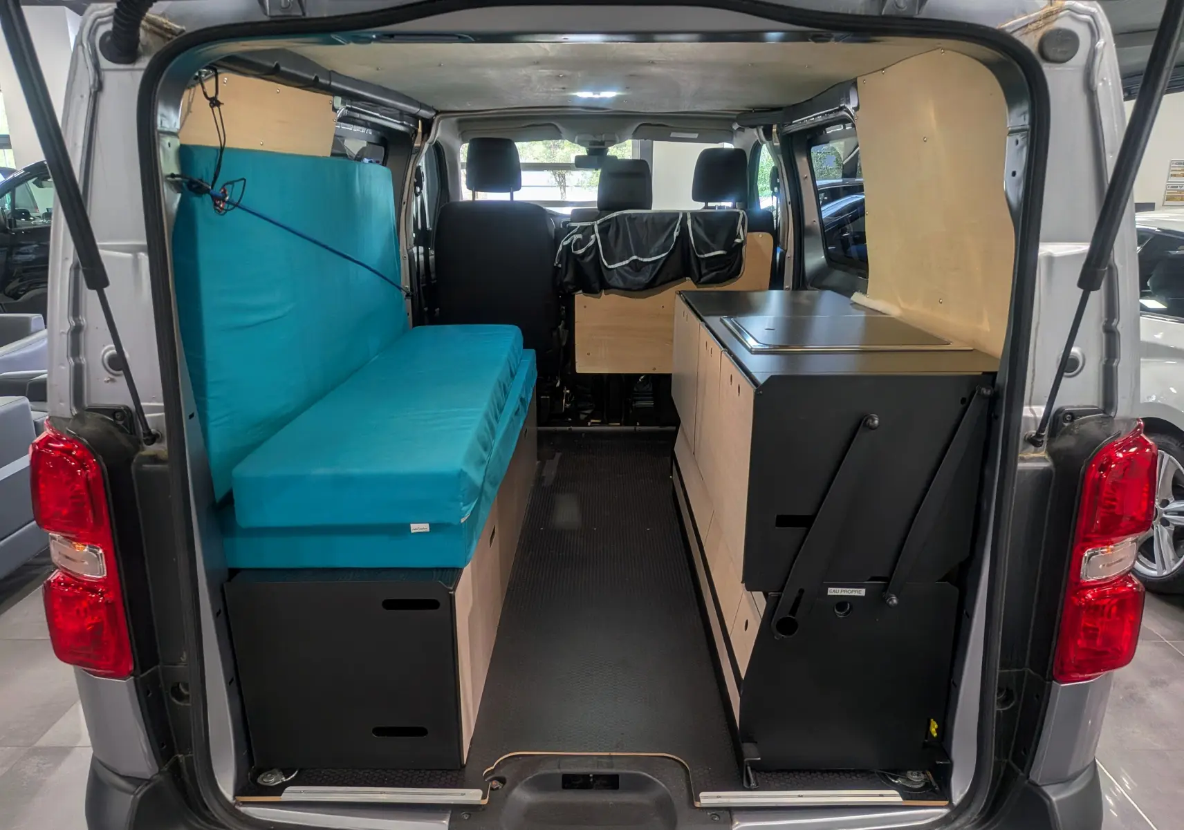 Vue arrière intérieure du Peugeot Expert Fourgon gris acier aménagé avec banquette bleue et mobilier bois intégré.
