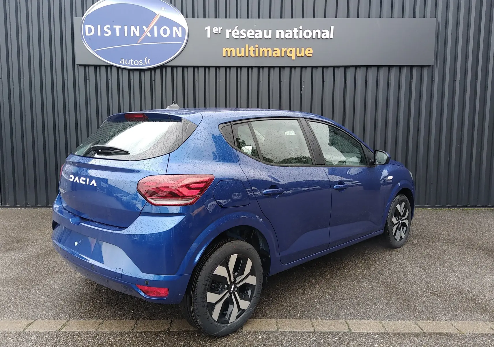 Vue 3/4 arrière droite d'une DACIA Sandero 2025 bleu iron avec jantes alliage et feux arrière LED sous pluie légère.