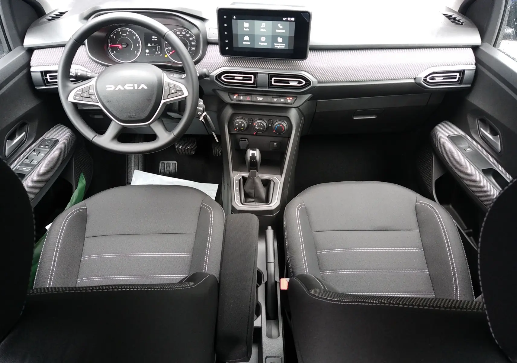 Vue intérieure frontale du tableau de bord et des sièges en tissu gris de la Dacia Sandero 2025 avec écran tactile et volant multifonction.