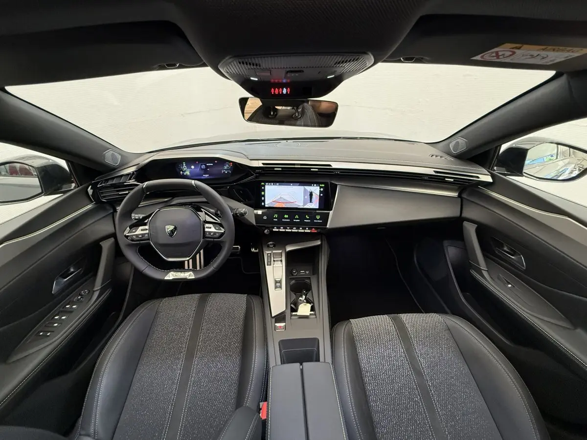 Vue intérieure avant de la Peugeot 308 Hybrid 136 e-DCS6 GT 2024, tableau de bord et sièges noirs avec volant cuir multifonctions.