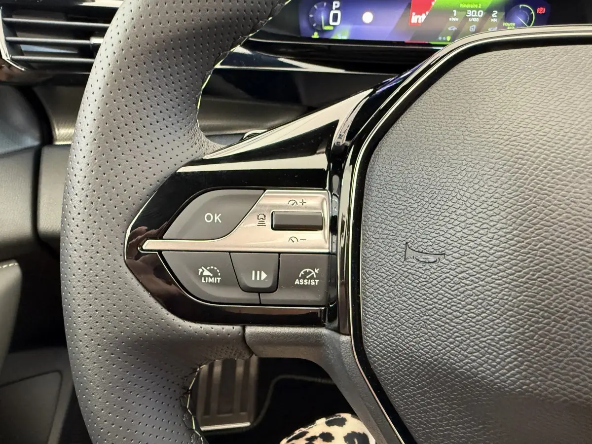 Gros plan sur le volant cuir perforé gris foncé de la Peugeot 308 Hybrid 136 e-DCS6 GT avec commandes multifonctions et tableau de bord numérique.