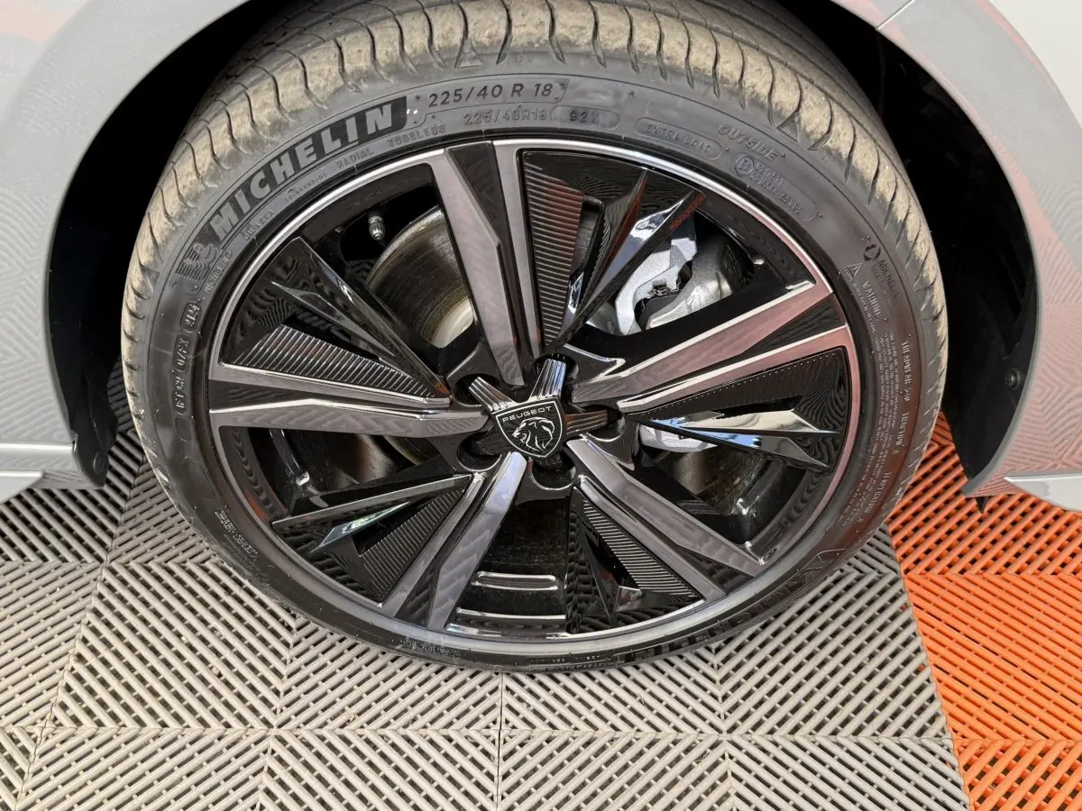 Gros plan sur la roue avant droite noire brillante d'une Peugeot 308 Hybrid 136 e-DCS6 GT gris foncé, avec logo lion central.