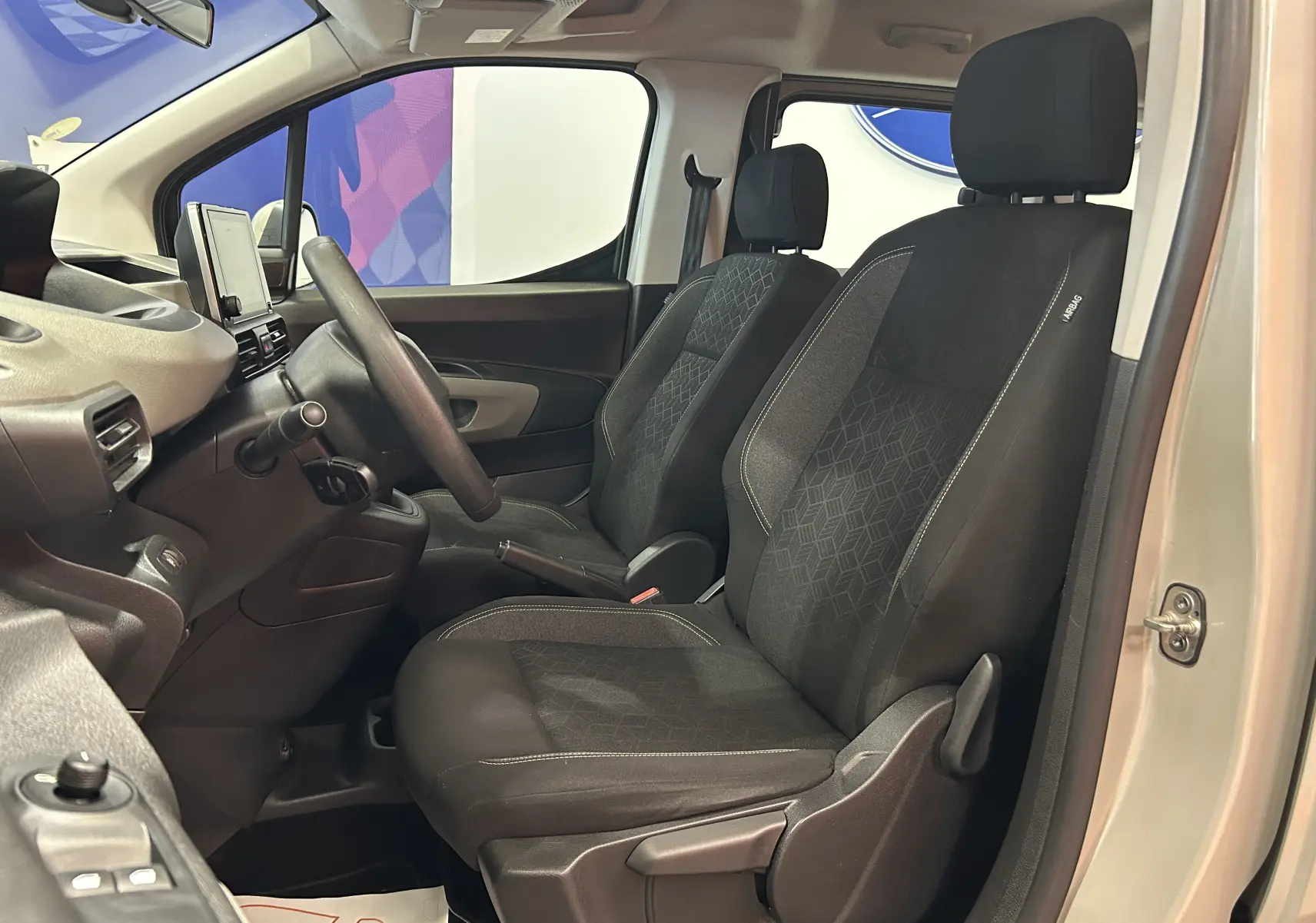 Vue intérieure côté conducteur du Peugeot Rifter gris sable 2019, sièges tissu avec motifs géométriques et tableau de bord moderne.