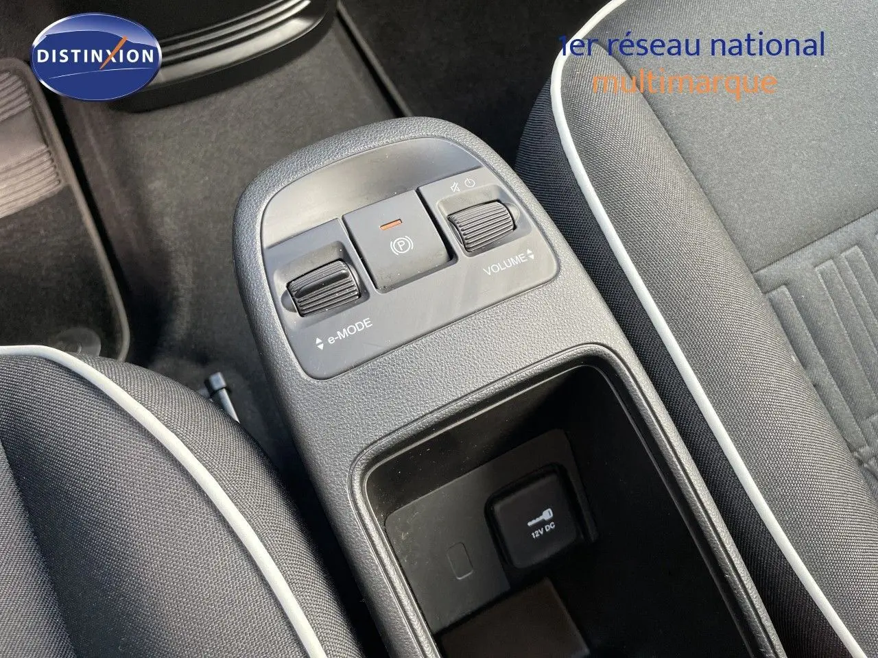 Gros plan sur la console centrale de la Fiat 500 électrique 2023, avec commandes e-Mode et volume, sièges tissu noir à liseré blanc.