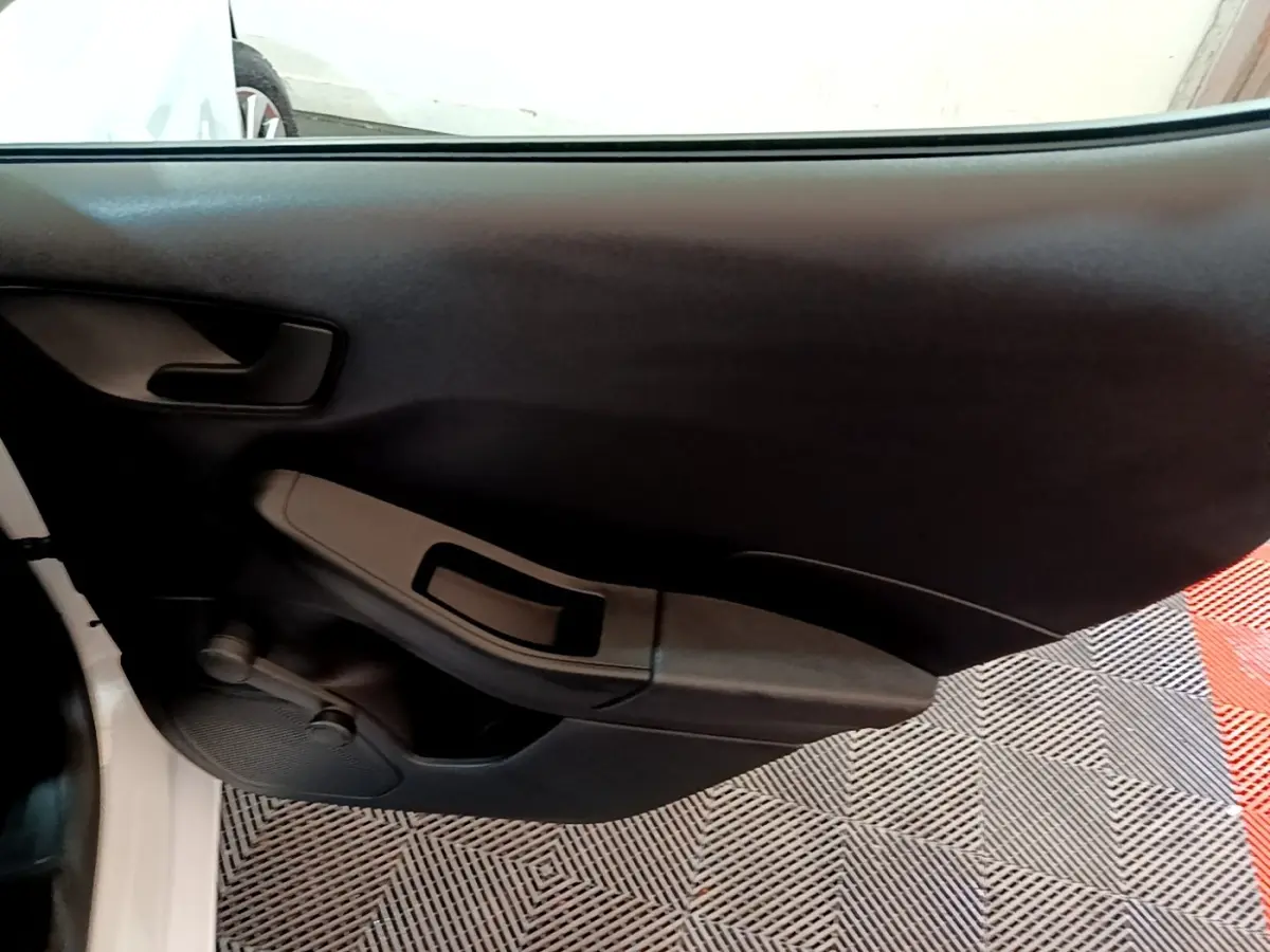 Vue intérieure sur la porte côté gauche d’une Ford Fiesta blanche, avec poignée et commande de vitre manuelle noire.