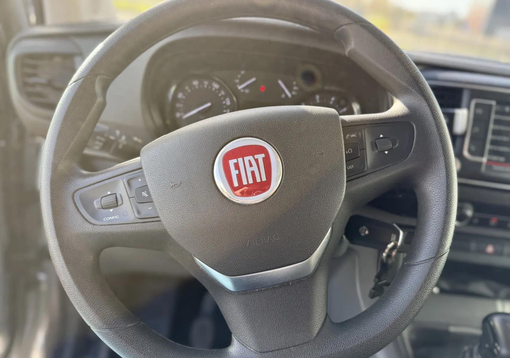 Vue rapprochée du volant noir avec logo FIAT rouge du Fiat Scudo Cabine Approfondie gris, tableau de bord en arrière-plan flou.