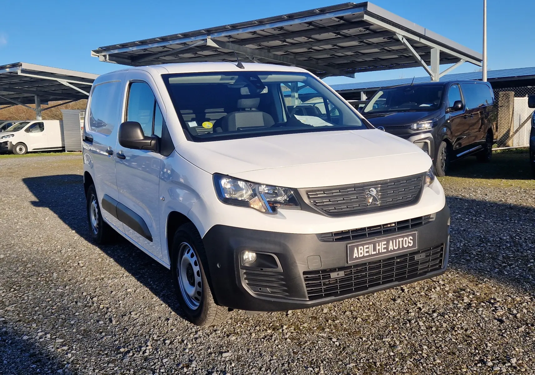 Vue 3/4 avant d'un Peugeot Partner blanc 2021 avec pare-chocs noir et plaque Abeilhe Autos sur un parking extérieur.