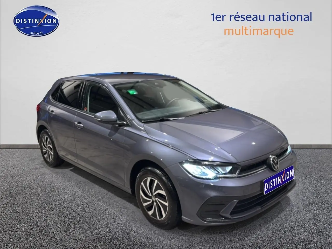 Volkswagen Polo gris cendre vue 3/4 avant droit avec phares allumés et jantes alliage.
