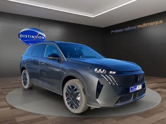 Vue 3/4 avant droite d’un Peugeot 5008 Hybrid gris titane avec jantes noires et calandre distinctive en intérieur showroom.