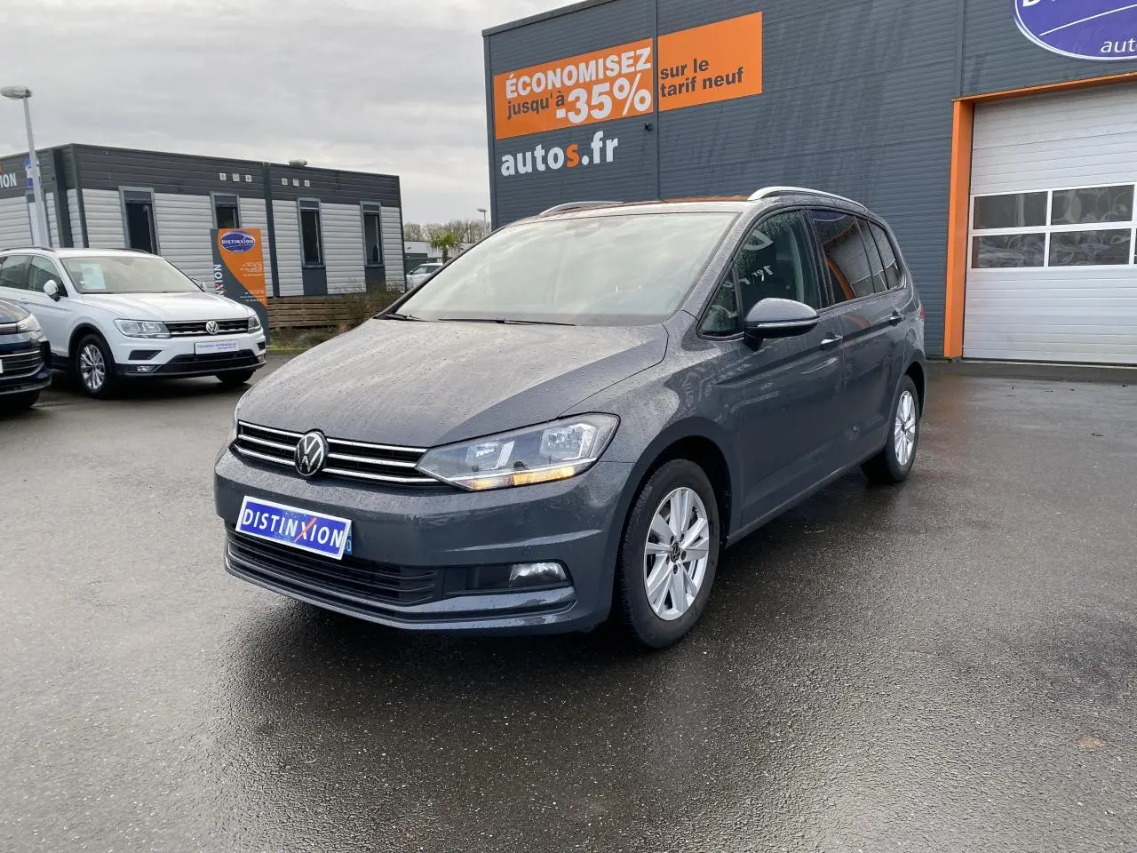 Volkswagen Touran gris dauphin en 3/4 avant droit sur parking devant un bâtiment commercial.