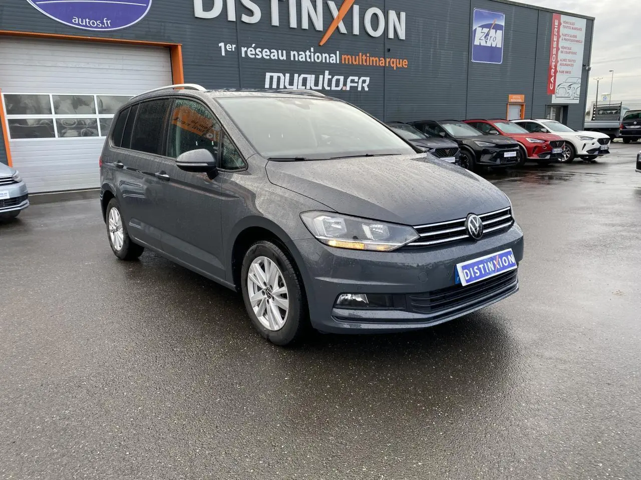 Volkswagen Touran gris dauphin vue 3/4 avant droit sur parking devant un bâtiment commercial.