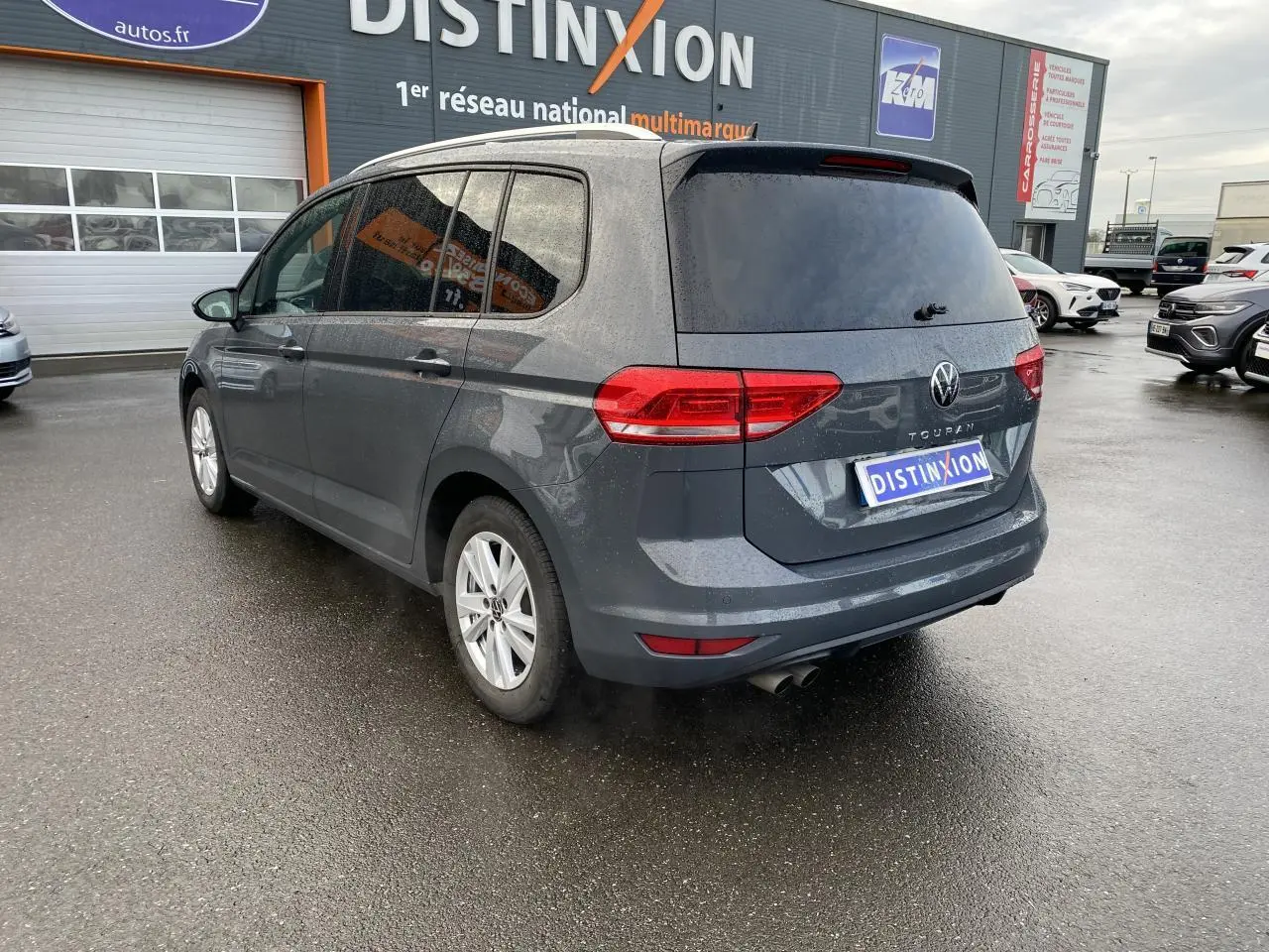 Vue 3/4 arrière droite d'un Volkswagen Touran gris dauphin avec vitres arrière surteintées, stationné devant un garage multimarque.