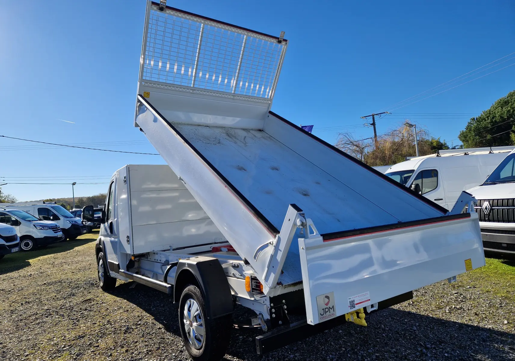 Vue 3/4 arrière droite d'un FIAT Ducato blanc avec benne acier relevée et grille de protection sur coffre.