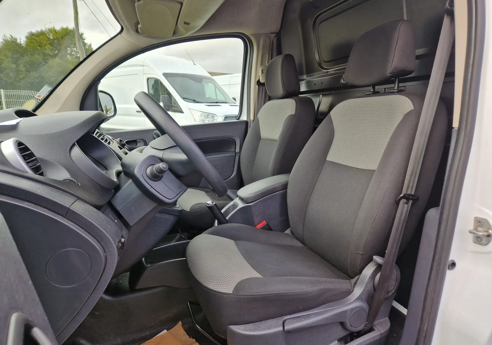 Vue intérieure côté conducteur du Renault Kangoo blanc 2020, montrant les sièges avant gris et la console centrale avec levier de vitesse manuel.